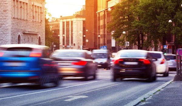 Cars travel along a city street lined with buildings and trees, with motion blur showing traffic movement.