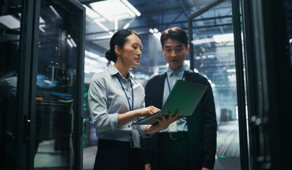 Two professionals working on a laptop while standing in a server rack corridor inside a modern data center.