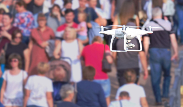 A white drone equipped with a camera hovers in front of a crowd of pedestrians.