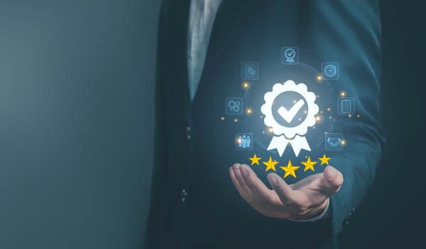 Person in a business suit holding an open hand beneath a glowing award ribbon icon with a checkmark, five gold stars, and surrounding business process icons.