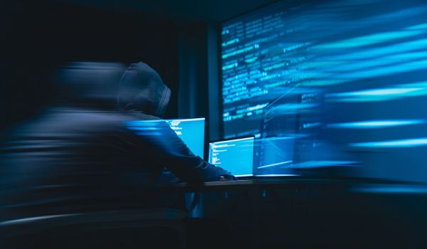 Hooded person working at multiple computer screens displaying code in a dark room.
