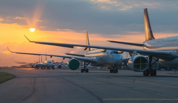 A line of commercial airplanes parked along a airport runway tarmac glowing beneath a bright golden sunset.