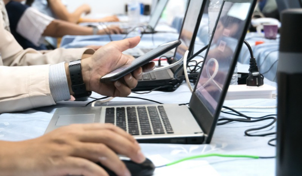 Close-up of a person holding a smartphone next to an open laptop at a desk lined with computers.