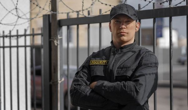 Security guard in a black uniform standing with arms crossed beside a metal fence topped with barbed wire.