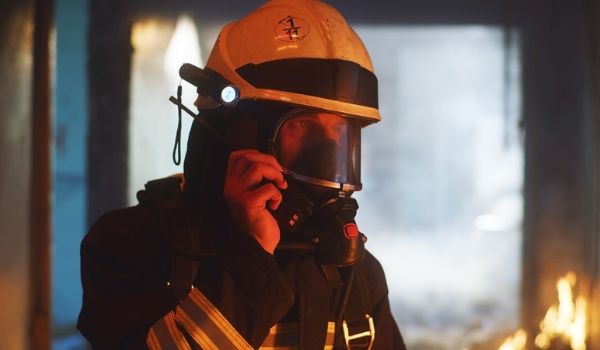 Firefighter wearing a helmet and breathing mask uses a handheld radio near flames inside a smoke-filled building.