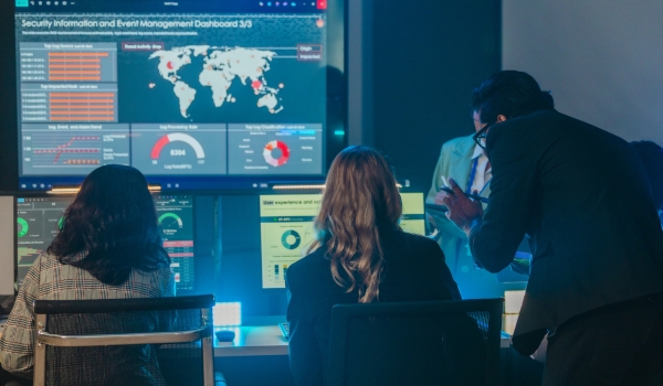 Security analysts monitor digital dashboards showing global map data and security metrics inside a dark security operations center.