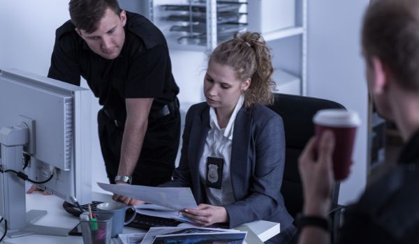 A seated investigator wearing a badge reviews documents with a uniformed colleague beside a computer monitor.