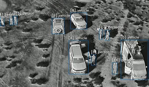 Grayscale aerial image of vehicles and people on a dirt road, with blue detection boxes labeling each person and vehicle.