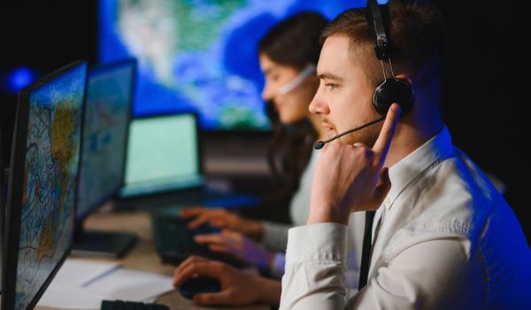 Two headset-wearing operators monitor computer screens displaying maps in a control room.