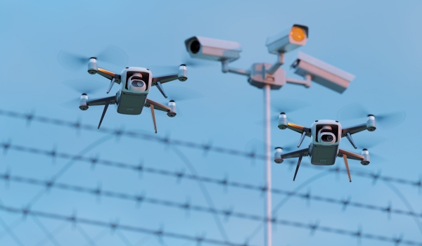 Two surveillance drones flying near a high-security fence topped with razor wire, with security cameras mounted on a pole in the background.