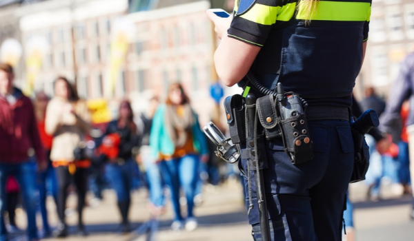 Uniformed officer with a holstered firearm stands in the foreground as pedestrians walk through a public area.
