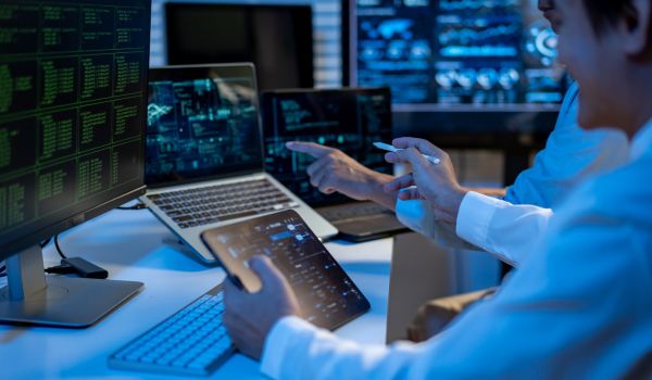 Cybersecurity specialist monitoring data on multiple screens in a dark office, using laptop and tablet to analyze code and network activity