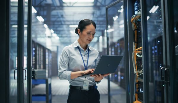 Asian Female System Administrator Using Laptop Computer for Software Configuration and Data Backup Monitoring While Standing Between Rows of Server Cabinets in AI Cloud Computing Center
