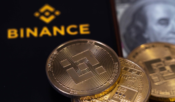 On screen mobile have logo of the binance trading website. And there are bnb coins and dollars laying nearby.